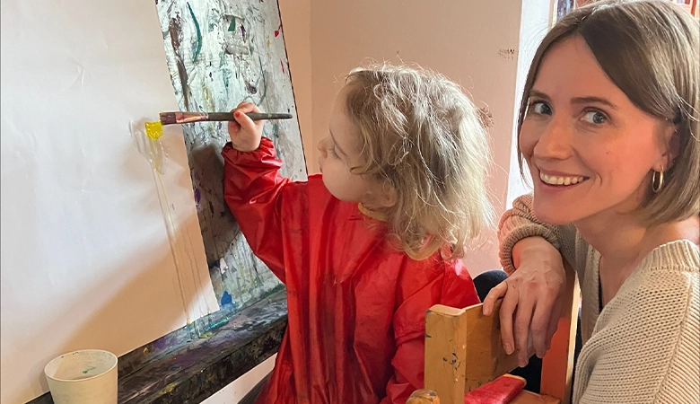 Kleines Mädchen malt mit einem Pinsel Farbe auf eine Wand. Eine Frau sitzt hinter ihr. 