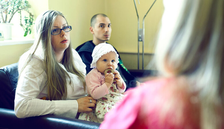 Eine Frau mit einem Baby auf dem Schoß und ein junger Mann sitzen auf einem Sofa. Sie blicken zu einer Frau, deren Rücken zu sehen ist.
